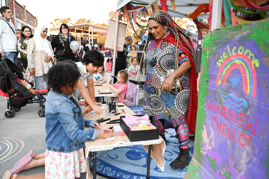 A woman in colourful dress interacts with children colouring at World of Cultures 2024